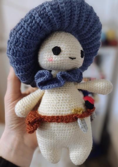 A crochet mushroom person. They are looking off to the right and have a little smile and blush on their cheeks. They have a pale cream head and body, with dumpy little eyes and legs. They have a blue ruff around their neck, and the gills of the cap on their head is also blue. The POV is slightly from below, so we can't see the top of the cap. They are wearing a brown tool belt, with a little bag, spade, and knife in it.