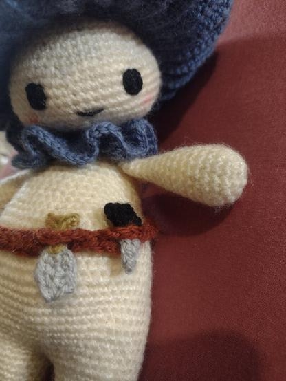 A crochet mushroom person looking down at the tool belt on their hips, specifically the spade and knife.. They are looking off to the right and have a little smile and blush on their cheeks. They have a pale cream head and body, with dumpy little eyes and legs. They have a blue ruff around their neck, and the gills of the cap on their head is also blue. The POV is slightly from below, so we can't see the top of the cap. They are wearing a brown tool belt, with a little bag, spade, and knife in it.