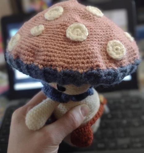 The top of a crochet muchroom person's cap. It is a dusty pink with lots of small cream circles on it, like an off-colour toadstool.