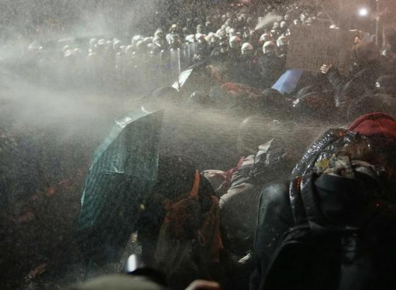 Das Foto zeigt eine Szene während der Demonstationen in der der Türkei um den 23.3.25.
Im Vordergrund ein paar Demonstrant:innen. In die Gruppe hinein schiesst aus der linken Seite des Bildes der Strahl eines Wasserwerfers. Die Demonstrant:innen versuchen sich mit Regenschirmen gegen den Strahl zu schützen.
Man sieht auch eine/n Demonstrant:in die/der ein Schild hochhält. 
Im Hintergrund sind sehr viel mit weissen Helmen und Schilden ausgerüstete Sicherheitskräft / Polizisten, die scheinbar(!) bereit stehen zum Einsatz. Sie schauen auf die kleine Gruppe Demonstrant:innen.