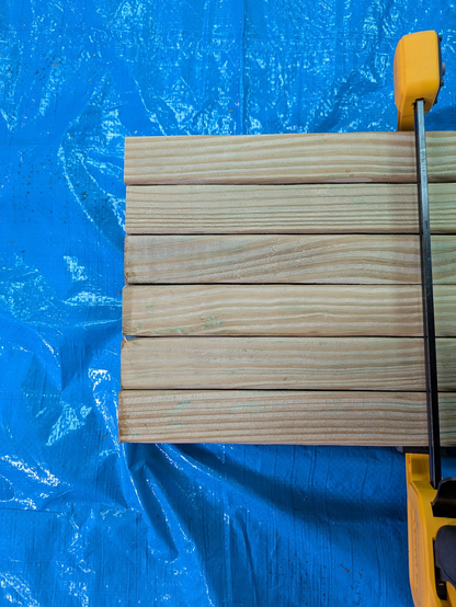 A photo of wood lined up side by side, with a yellow clamp holding them together, against a blue tarp. The ends of the boards are nearly flush.