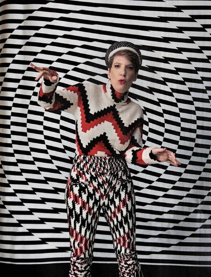 A person wearing a geometrically patterned outfit with red, black, and white zigzag designs on both the sweater and pants. They are posing with outstretched arms against a swirling black and white checkered background that creates an optical illusion effect. They have short hair and are wearing red lipstick.