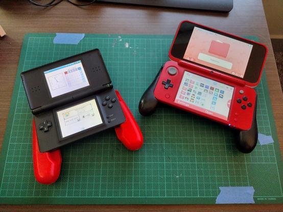 A picture showing a black DS Lite with a red 3D printed grip, next to a red 2DSXL with a black grip.