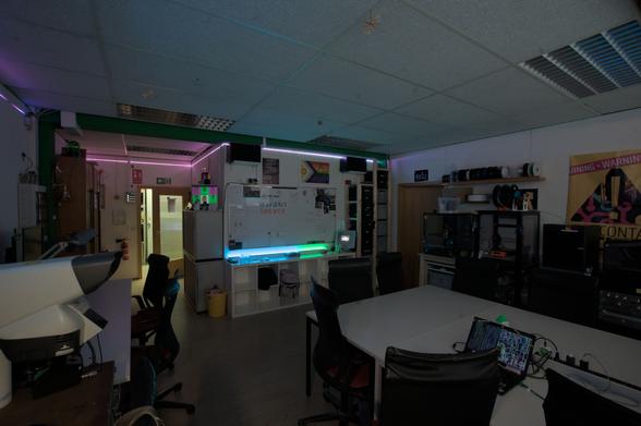 Ein weitwinkliges Bild von dem Labor im vspace. Der Raum ist gefüllt mit verschiedenster Ausstattung wie etwa unseren 3D-Druckern mit Regalen voll mit Filament darüber, Tischen mit dazgehörigen Stühlen, Plakaten sowie ein Whiteboard mit Zeichnungen und Postkarten, ein Statusdisplay auf einem Schrank und ein RGB-Streifen, der sich an der Wand entlang zieht. Auf einem Quer aufgestellten Kallax-Regal unter dem Whiteboard steht ein 1D-Pong und es ist ein Laptop auf dem Tisch zu sehen.