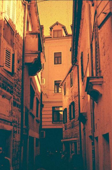 A portrait orientated redscale photo of a narrow street. The bottom features dark, hard to distinguish silhouettes of people, while the top of the frame featuring the tops of buildings has much better lighting