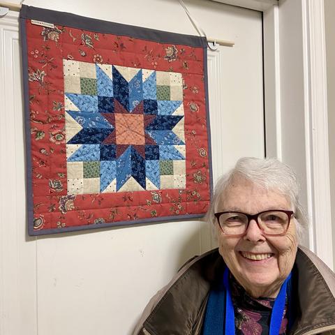 A small wall quilt featuring a flower pieced from blue and purple fabric on a cream background. The flower center is a pink square. The dark blue fabric breaks the flower pattern into a pinwheel (I wish I'd stuck to mid-values). There is a wide border in a salmon pink dutch heritage floral print, the same as I used on the reverse.