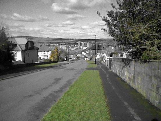 A quiet suburban street slopes gently downward, leading to a distant cityscape nestled between rolling hills. The image is selectively colorized, with vibrant green grass contrasting against the monochrome surroundings. A yellow-tinged bush stands out near the roadside, likely due to overlapping hues in the filtering process. The sky is dotted with fluffy clouds, casting soft shadows over the landscape. A sidewalk and a stone wall run alongside the road, adding depth and perspective. Houses with pitched roofs line the street, their neutral tones blending into the grayscale effect. The scene captures a peaceful moment in an urban-rural transition.