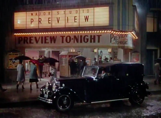 An exterior shot of an old theater on a rainy evening. An old-timey car is parked outside, and people walk in holding umbrellas. The marquee reads, "Major studio preview" and "Preview tonight".