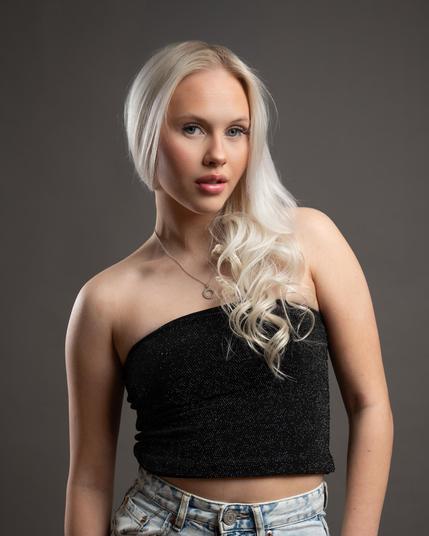 The image shows a person with long, light blonde hair wearing a black, sparkly off-the-shoulder top and light denim jeans. The background is neutral, likely used to emphasize the subject. The person is also wearing a necklace.