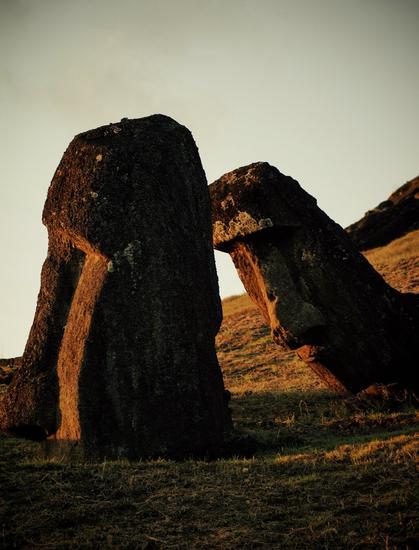 Sunset on Rapa Nui. A close up view of two moai. The one on the right is leaning forward and, thanks to the angle, seems to be leaning on a more upright statue (though this one is buried in the ground up to the tip of its nose).