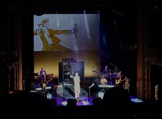 Vorn der Sänger und die band. Darüber sieht man in einer Live Zeichnung david Bowie in einem gelben Anzug mit Mikrofon