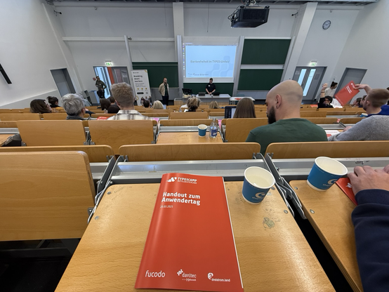 Ein Blick in den Hörsaal beim TYPO3 Anwendertag 2025 in Jena – vorne läuft gerade eine Session zu Barrierefreiheit im TYPO3-Umfeld, das Handout liegt griffbereit. Tolle Atmosphäre und geballtes Wissen!