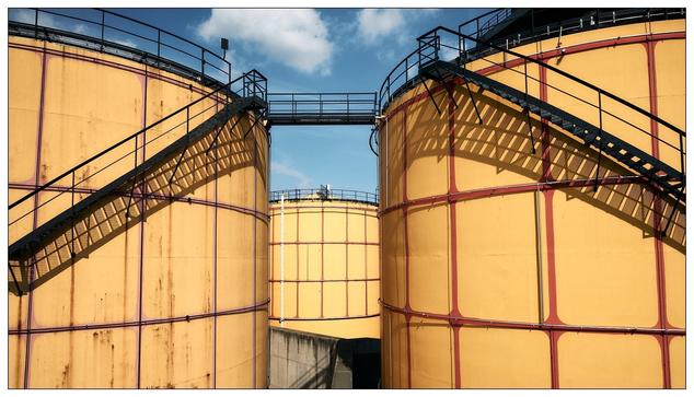The image shows a set of large industrial storage tanks, typically used for holding liquids such as water, chemicals, or oil. These tanks are yellow with red structural elements such as support beams and stairs. There are external staircases wrapping around the tanks, likely for maintenance access. The scene includes a backdrop of a blue sky with light clouds, suggesting a clear, sunny day.
