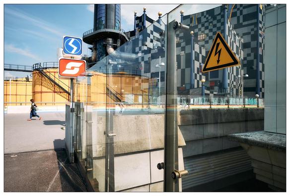 This image features a complex urban scene with multiple reflective surfaces, creating a visually intriguing overlay of elements. This creates a nearly collage-like effect. Specifically, it includes:

1. **Road signs:** There are multiple traffic signs visible including a direction arrow, a speed limit sign, and an electric hazard warning sign.
2. **Industrial Background:** In the background, there appears to be an industrial setting with structures such as a tall chimney stack and yellow scaffolding.
3. **Glass Reflections:** The foreground prominently displays a glass surface reflecting various elements like the road signs, blending them with the actual physical objects behind the glass.
4. **A person:** An individual is visible in the mid-ground, moving through the space on foot, wearing red shoes, adding a sense of everyday human activity to the scene.
5. **Architectural Details:** The photo includes modern architectural elements like sleek glass panels and a stairway, suggesting an urban environment with contemporary design.

Overall, the photo captures a dynamic urban environment where industrial, architectural, and everyday human elements intermingle, highlighted through an artistic use of reflection and perspective.
