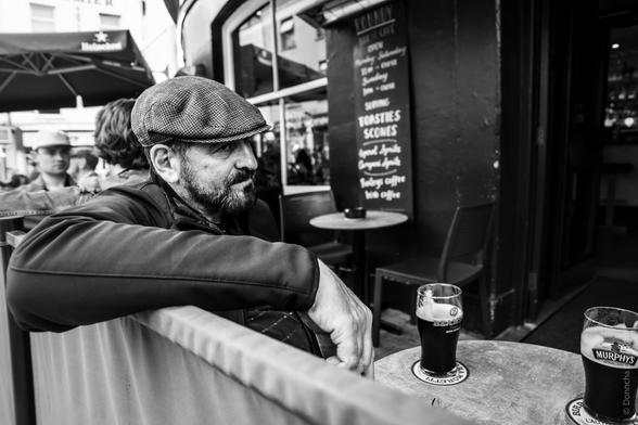 A black-and-white photograph of a man wearing a flat cap and jacket, sitting outdoors at a pub table in Cork, Ireland. He leans on a barrier, with two pints of Murphy's stout on the table in front of him. A chalkboard menu behind him lists items such as "Toasties" and "Scones" alongside coffee options. Other patrons are visible in the background, seated under umbrellas.