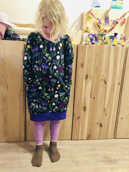 A white 5 year old girl standing in front of light wooden 80 cm high cabinets and standing on a light wooden floor, wearing brown socks, lavender coloured leggings and her new dress, described in more detail in the next photo’s ALT. She’s looking down, her blond half long hair obscuring her face, hands in the pockets of the dress. On one of the cabinets in the background is the seasonal table from previous posts.