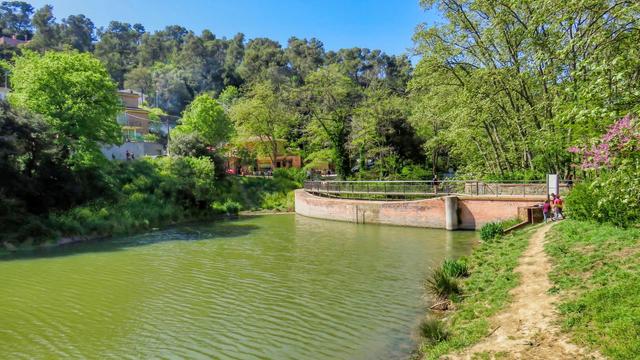 Aquesta imatge mostra un estany tranquil o llac envoltat d'exuberant vegetació. Un corriol corre al llarg de la vora de l'aigua, mentre que un pont de maó condueix a una àrea més desenvolupada que sembla incloure un restaurant o un altre establiment. Les cases i la vegetació més densa s'eleven al fons, indicant un entorn natural i residencial.