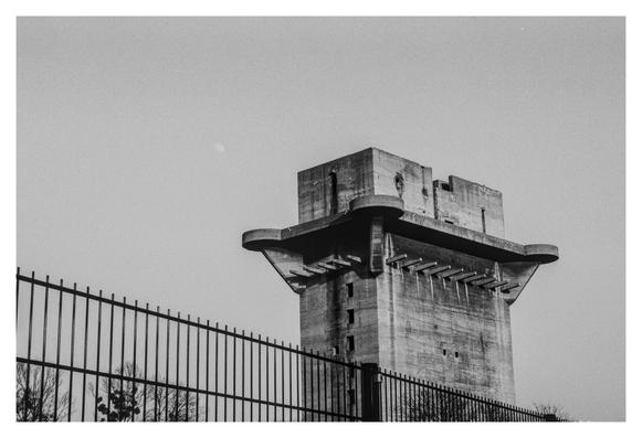 Black and white photograph of the L-tower of the flak tower complex in Augartenpark. In the foreground, the top of a fence slopes down from the left. The moon, which was visible in the daytime sky, can be seen to the left above the L-tower.