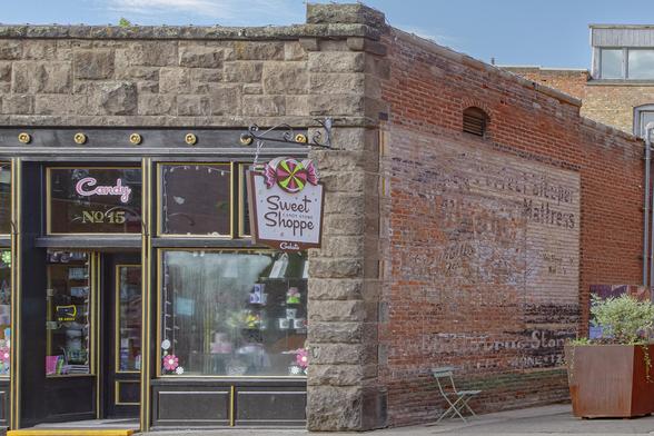 Sweet Shop Sweet Spot - Candy, sweets, gelato and gifts in an old downtown Flagstaff shop with the old limestone and red brick exterior. The windows are fogged with moisture and it was going to soon rain again. Alley side view shows an old sign painted and fading off the bricks, for a mattress sold long ago. The entrance is old fashioned with black painted wood, gold painted trim around door and window panels and it is dark still, early morning.