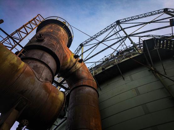 Color photo of an old industrail site. Part of the photo is taken up by the side of a gasholder, Part by some rusted pipes with a very large diameter. The POV is upwards, with the sky as backround.