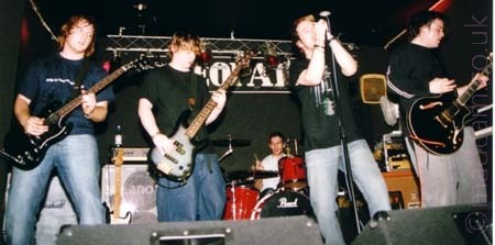 5 men standing on a stage, all members of a band.
On the left is a man with long brown hair, wearing a blue t-shirt, blue jeans, and wielding a black guitar.
Next to him is a man with long brown hair, wearing a black t-shirt and blue jeans, playing a black and grey bass guitar.
At the back, a man with short dark hair and wearing a grey t-shirt is sitting behind a red and chrome drum kit.
Next is a man with short brown hair, wearing a black t-shirt and pale blue trousers.
He is holding a microphone mounted on a stand in both hands to his mouth, singing in to it.
On the right is a man with short dark hair, wearing a black sweater and grey trousers, playing a black electric guitar.
The walls and ceiling behind them are black, with a metal lighting rig running across the top of the back wall.