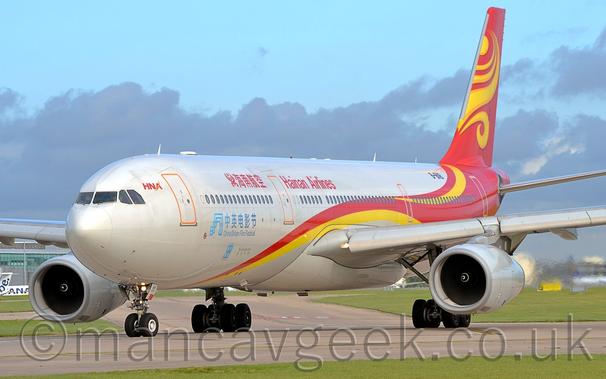 Side view of a white, twin engined jet airliner taxiing from right to left.
The plane has a red and yellow stripe running from under the lower forward fuselage, swooping over the wings, and up into the tail, which is almost entirely red, with a swirling yellow design.
There are red "Hainan Airlines" on the upper forward fuselage, in a Chinese script in front of the mid-cabin door, and in English aft of that door.
There are blue "China Britain Film Festival" titles on the lower forward fuselage, in a Chinese script under the cabin windows, and in English below that.
The registration is on the upper rear fuselage, in black.
A strip of lush green grass at the bottom of the screen marks the edge of the taxiway, with more grass delineating taxiways in the distance, visible under the planes body.
Pale blue skies and purple clouds fill the rest of the frame in the background.