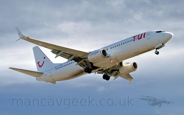 Side view of a white, twin engined jet airliner flying from left to right, with undercarriage lowered and flaps extended from the rear of the wings, suggesting it is about to land..
The plane has red "TUI" titles on the upper forward fuselage, and a red logo on that tail that appears to be a mix of the letters "T", "U", and "I", and a winking smiley face.
There are blue "TUI[dot]com" titles on the sides of the white engine pods.
The registration "G-TUKR" is visible on the upper rear fuselage, next to a small British flag.
Cloudy grey sky with patches of blue fills the rest of the frame.