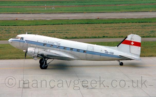 Side view of a World War 2-era twin propellor airliner taxiing from right to left.
The plane is white, with a grey belly, and a pale blue stripe running along the body.
There are dark blue "Classic Air" titles on the upper fuselage, with the registration "HB-ISB" on the upper rear in black.
There is a red band around the tail with a white cross in the middle.
Grey tarmac apron fills the foreground, with grass in the background leading up to a runway, and more grass beyond that filling the rest of the frame.