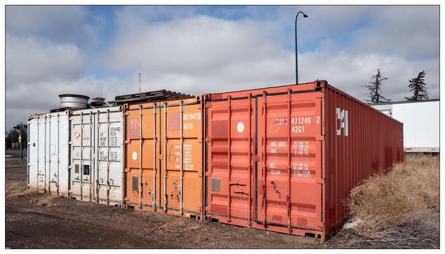 A collection of industrial cargo containers. These containers, typically used for shipping goods over long distances via road, sea, or rail, appear to be placed in a storage or depot area. The containers are of various colors, primarily rust red and beige, and have different numbers and coding on them, which indicates their identification and tracking details. The environment suggests a dry, possibly semi-arid area, as evidenced by the surrounding dry grass and the sky which appears partly cloudy.