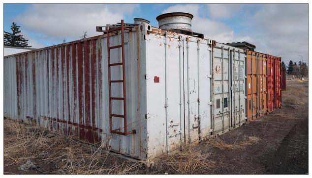 A line of weathered shipping containers. These containers appear old and rusted with varying colors, primarily white and red. Some have visible doors, numbers, and markings. The scene is set in an outdoor area with sparse grass and a clear sky, suggesting a storage or unused area possibly on the outskirts of a town or in an industrial zone.