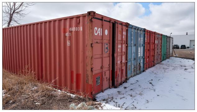 Arow of intermodal shipping containers, which are standardized metal containers used to transport goods securely by ship, rail, and road. The containers in the image are of various colors, predominantly red and blue, each marked with different identification codes and logos. The ground is partially covered with snow, indicating it's taken in a colder season or climate. The background features a leafless tree and a building, suggesting the containers are stored in an industrial or commercial area.