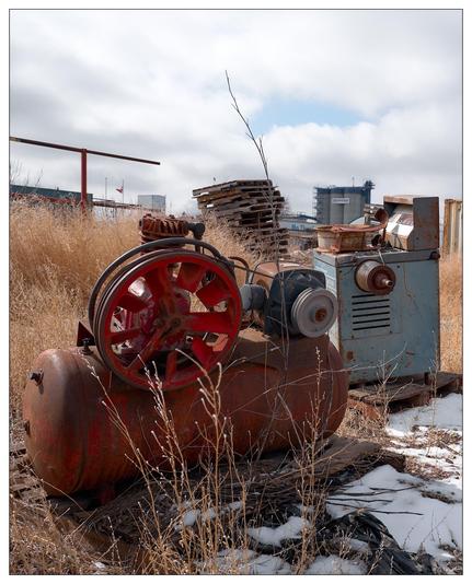 A scene of industrial decay featuring two pieces of old machinery in a state of disrepair. In the foreground, there's an air compressor with a red wheel and a rusty, red tank. Beside it is another piece of aged industrial equipment in blue. Both are surrounded by dry, brown grass and patches of snow. In the background, a stack of wooden pallets and some industrial buildings can be seen under a cloudy sky, suggesting a larger industrial area likely past its prime.