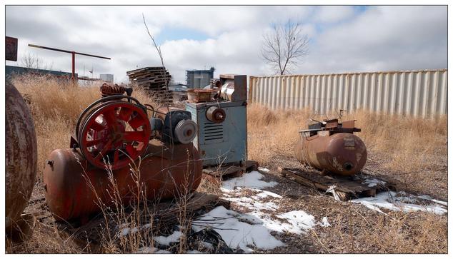 A scene of industrial decay, featuring discarded and rusty machinery in a field. There are several prominent objects including a large red air compressor, another machine with a blue and grey body, and a spherical metal tank with rust and deterioration evident on its surface. The environment is somewhat barren, with sparse vegetation, patches of snow on the ground, and a cloudy sky. The backdrop includes a metal fence and industrial structures that might be part of a factory or similar facility. The overall scene evokes a sense of abandonment and disuse.