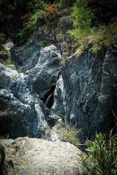 The image depicts a rugged, rocky landscape with a prominent gorge. The foreground features a large, smooth rock with a rough texture, surrounded by smaller rocks and sparse vegetation, including a bush with pink flowers. The middle ground showcases the gorge, with steep, columnar rock formations that are dark gray and vertically striated, suggesting volcanic activity. The gorge appears to be a natural water channel, with a visible stream or waterfall at the bottom, partially obscured by the rock walls. The background is filled with dense greenery, including trees and shrubs, with some foliage showing autumnal colors. The lighting suggests a sunny day, with shadows cast by the rock formations, enhancing the depth and texture of the scene.

Provided by @altbot, generated privately and locally using Ovis2-8B