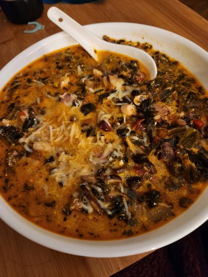 A while bowl filled with dark stew. The broth is tomato based, with some dark greens and yellow cheese on top. A white ceramic soup spoon is in the bowl.