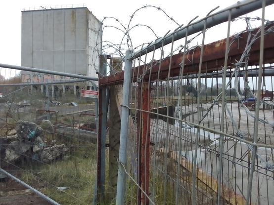 Another view of the building closest to the wharf, the hoppers of its silos are visible at ground level through the RC post structure. The wharf and the gutway of the river at low water are also visible though obscured by the remains of RC and rusted steel fencing in front of which is barbed wire topped Heras fencing