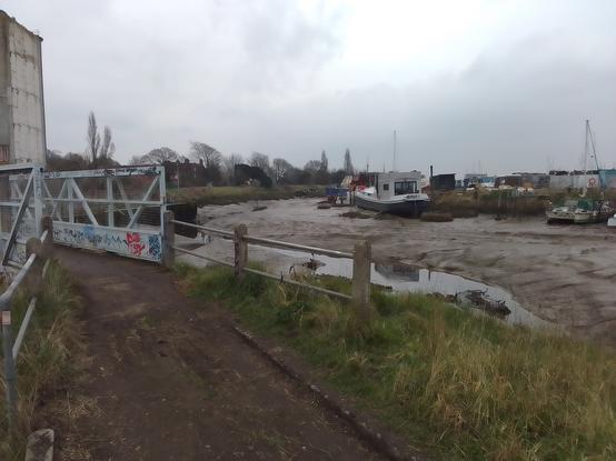 A view of the ned of  a bridge at the head of the navigation, with the silo building and the wharf, the gutway and mudbanks of the river. On the other side, a boatyard with various craft moored.