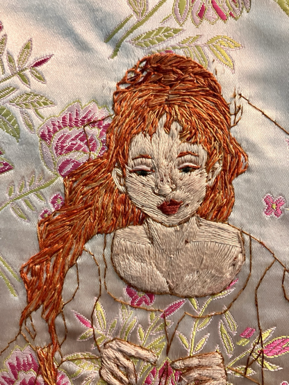 An embroidered portrait in progress of a pale woman with bright copper hair, looking down toward her knitting hands.