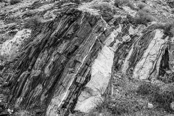 The image depicts a rocky landscape with prominent stratified rock formations. The rocks exhibit distinct horizontal layers, with varying shades of gray and white, indicating different mineral compositions or sedimentary processes. The foreground features a large, inclined rock face with visible striations, suggesting geological activity over time. The background shows additional rock formations and sparse vegetation, including small bushes and grasses, typical of a dry, arid environment. The black and white color scheme emphasizes the textures and contrasts within the rocks, highlighting the ruggedness of the terrain.

Provided by @altbot, generated privately and locally using Ovis2-8B