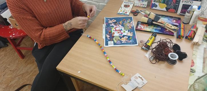 Foto von einer Person, die an einem Tisch sitzt und ein Armband auffädelt. Auf dem Tisch liegen viele Bastelmaterialien und ein queeres Ausmalbuch