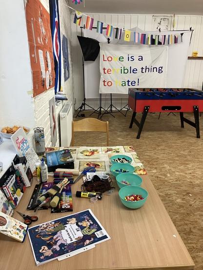 Foto von einem Raum: vorne ist ein Tisch mit vielen Bastelmaterialien zu sehen. Im Hintergrund steht ein Tichkicker. Darüber hängt eine Wimpekette mit vielen queeren Flaggen sowie ein Banner mit der Aufschrift "Love is a terrible thing to hate"