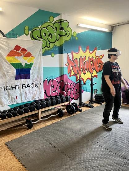 Foto von einer Person mit einer VR-Brille. Im Hintergrunf hängt ein Banner, auf dem eine Faust in Regenbogenfarben sowie eine AIDS-Schleife mit der Aufschrift "Fight back" abgebildet sind.