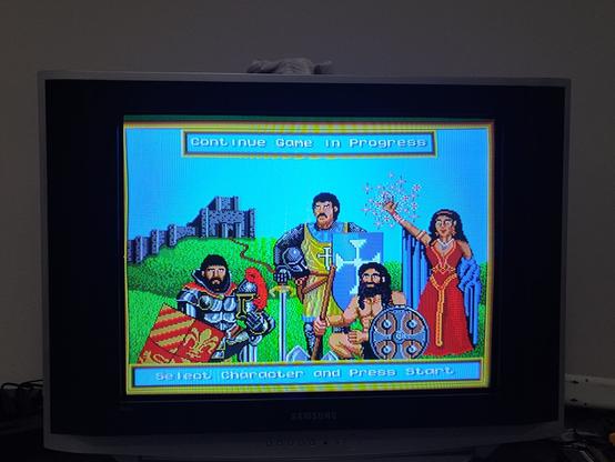 The image shows a retro video game character selection screen displayed on a CRT television. The game is King's Bounty in the Genesis, featuring four characters: a knight in silver armor with a red cape, a warrior with a shield and axe, a bearded man in a tunic holding a staff, and a sorceress in a red dress casting magic. The background includes a green landscape with a castle in ruins. The text on the screen says "Continue Game in Progress" at the top and "Select Character and Press Start" at the bottom.

Português:
A imagem mostra uma tela de seleção de personagens de um jogo retrô exibida em uma televisão CRT. O jogo é , King's Bounty no Genesis, com quatro personagens: um cavaleiro de armadura prateada com capa vermelha, um guerreiro com escudo e machado, um homem barbudo vestindo uma túnica e segurando um cajado, e uma feiticeira de vestido vermelho conjurando ma