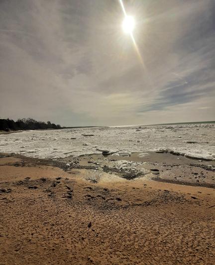 An ice strewn lake shoreline with a sand and stone beach foreground, a white soup  of ice chunks near shore, stretching  out to an evergreen shoreline to the left, and blue open water at the horizon, where it meets a light  blue sky filled  with wispy clouds framing and filtering the sun at the centre-top of the image.
