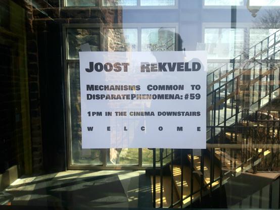 A small poster on a glass door, saying "Joost Rekveld, Mechanisms Common to Disparate Phenomena; #59, 1 pm in the cinema downstairs, welcome"