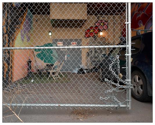 A gated area, partially enclosed by a chain-link fence gate with a large hole on the right hand side.   There is a metal chain and lock looped through the chain-link and around the end of the gate, making a rough repair to prevent people going through the hole.  Through the gate, you can see a small outdoor seating area with chairs, the side of a building, a portion of a car, a colorful artwork on the wall. The setting appears to be somewhat industrial or utilitarian, possibly a back alley or service area behind buildings.