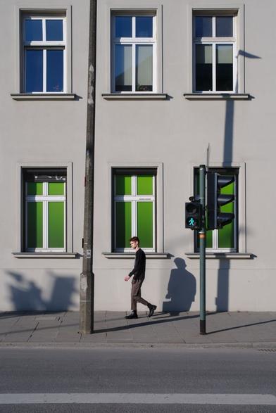 Man walking along housefront on which the sun casts sharp shadows. Three windows with green screens and a green traffic light n structure the frame.