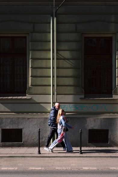 Couple walking along greenish housefront stark spring sun.