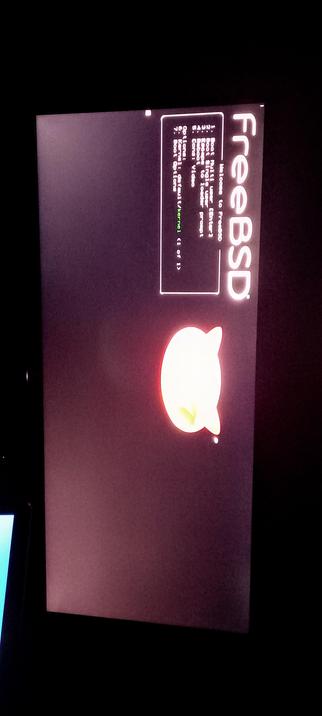 The photograph shows a IPS LED panel displaying the boot menu of the FreeBSD operating system. The screen is predominantly dark with a red logo in the center, which is the FreeBSD mascot, a stylized cat. On the right side, the text "FreeBSD" is displayed vertically in white. The boot menu is presented in a white box with a black background, listing various boot options such as "boot," "boot kernel," and "boot kernel text." The text is in white, with some options highlighted in green. The screen is framed by a black border, and the background is dark, emphasizing the text and logo.

The image was captured with a telephone sensor which clearly shows the horrific low Quality of the Light vs the dark.

Phones fail when shooting in darkness