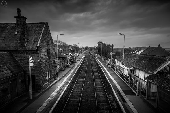 Monochrome shot over railway lines leading into the distance. Station house and platform in shot, heavy sky above